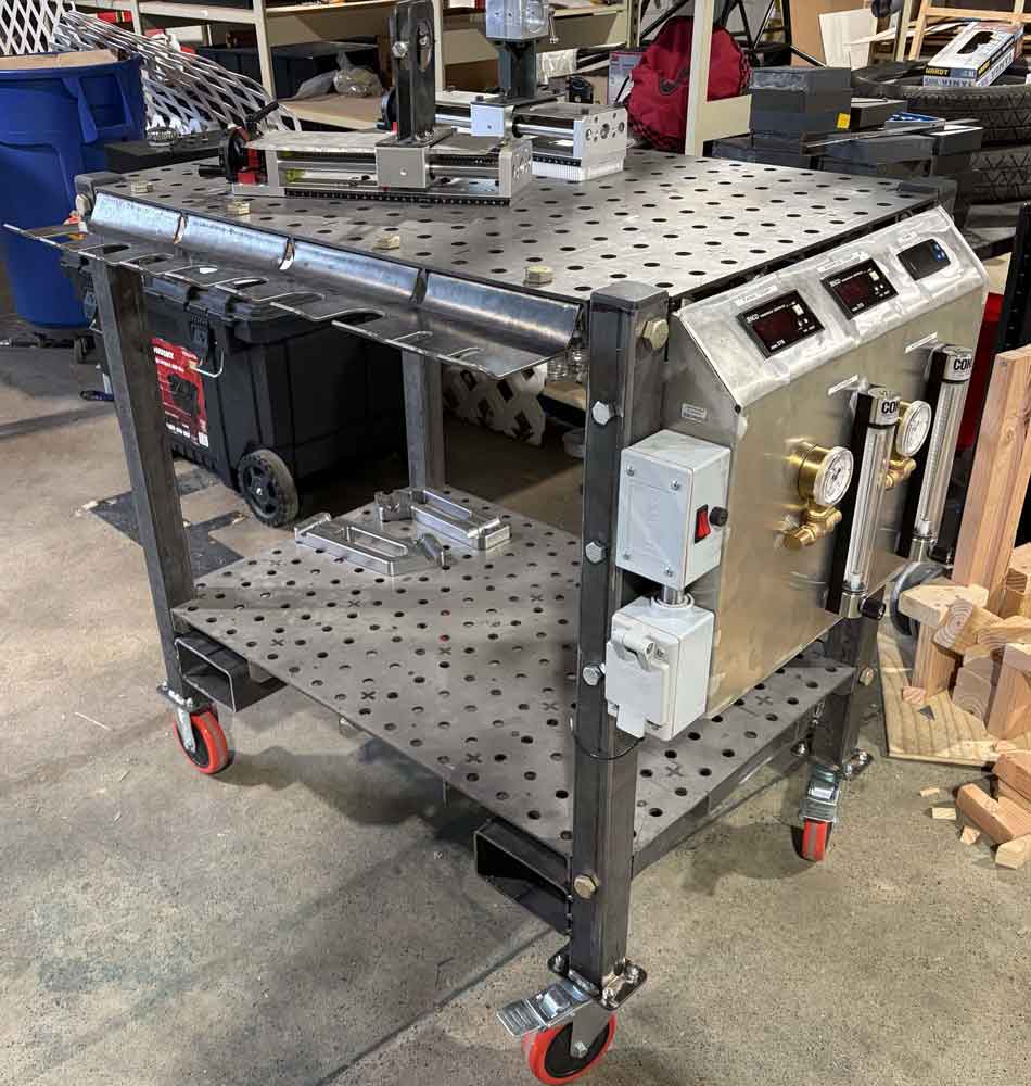 Photograph of the complete table subsystem showing welding tabletops, mounted components, sample holder rack, water reservoir, wheels, and fire extinguisher